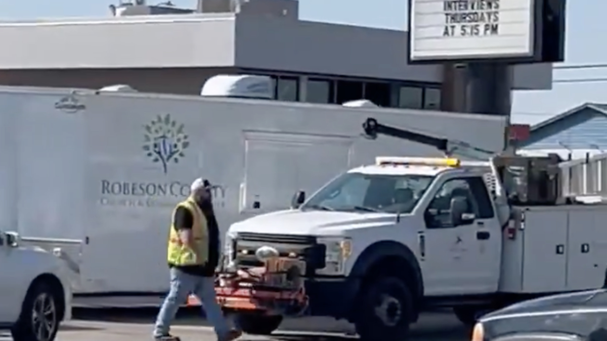 Person in high visibility vest walking across a parking lot, with trucks and trailers in the background