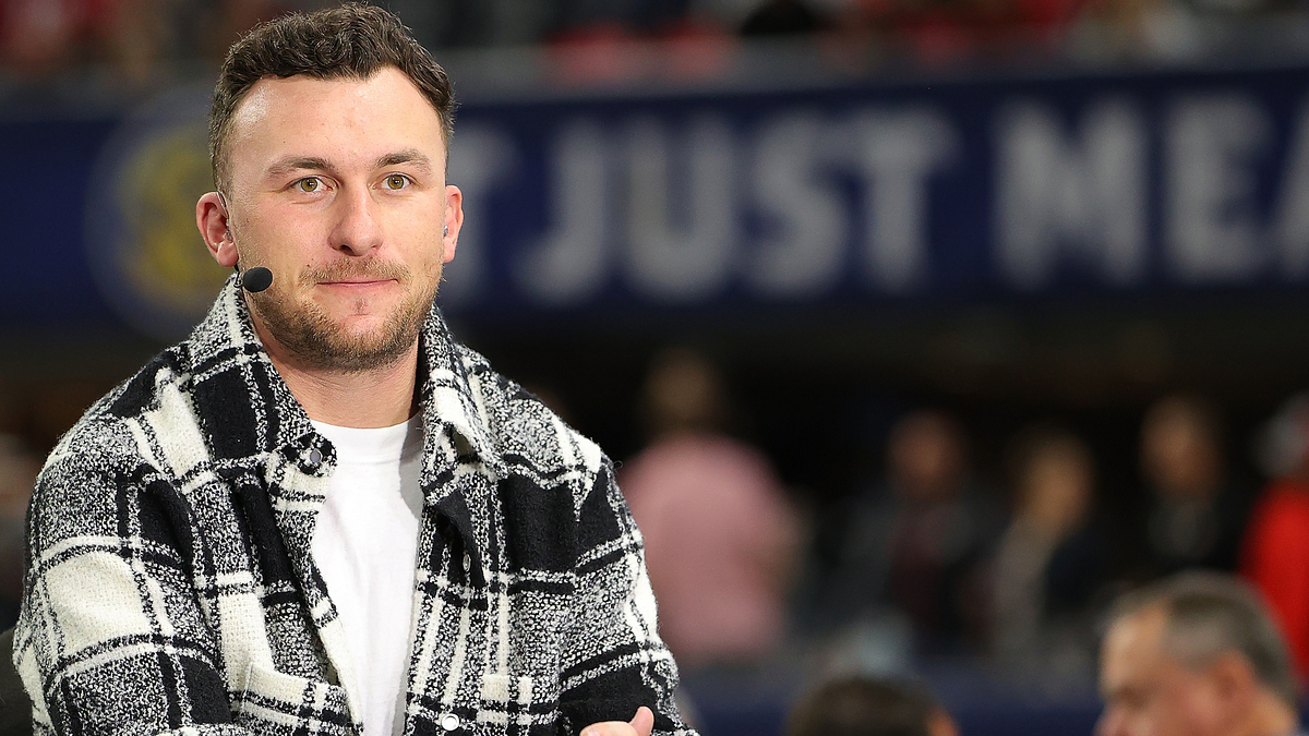 Person wearing patterned jacket at a sports event, with microphone and earpiece, seated, with blurred audience in background