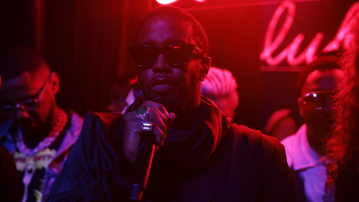 A musician in sunglasses performs with a microphone, other artists in the background onstage