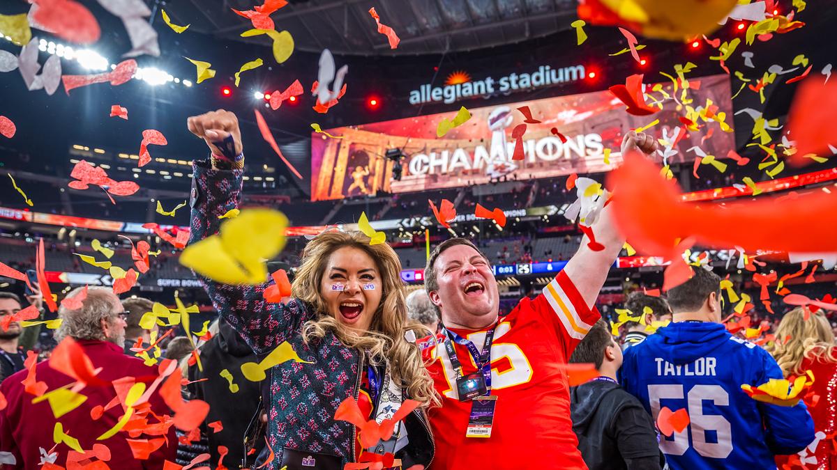 Two excited fans celebrate amidst falling confetti at a stadium with a "CHAMPIONS" sign in the background
