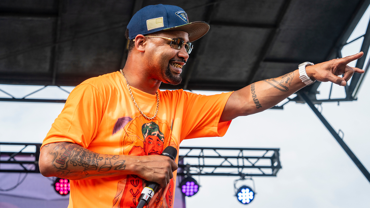 Juvenile on stage performing with microphone, wearing an orange shirt and a blue cap