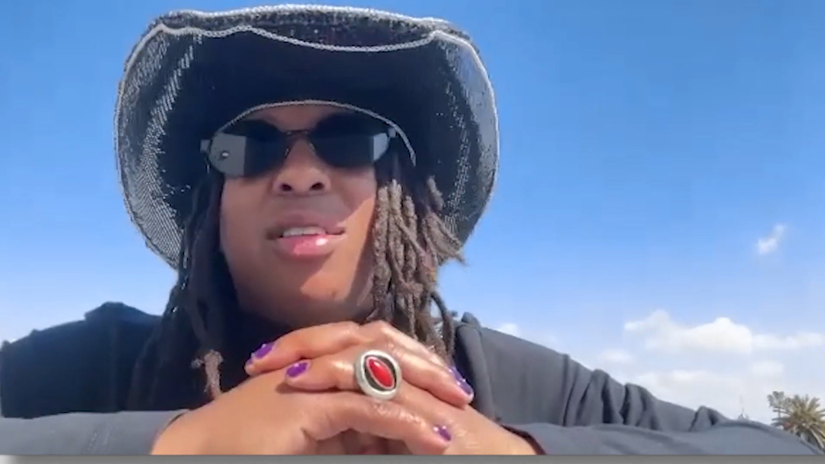 Person with sunglasses and a wide-brimmed hat, hands clasped in front, clear sky behind