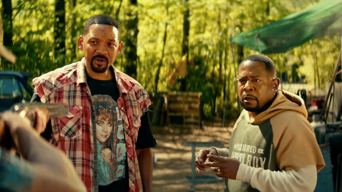 Two actors in a forest scene, one in a flannel shirt, the other in a hoodie and jacket. They appear in a tense conversation