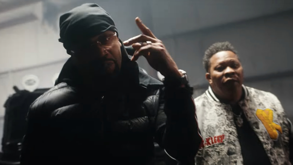 Two hip-hop artists in a moody video set, one making a hand gesture, the other in a patterned jacket