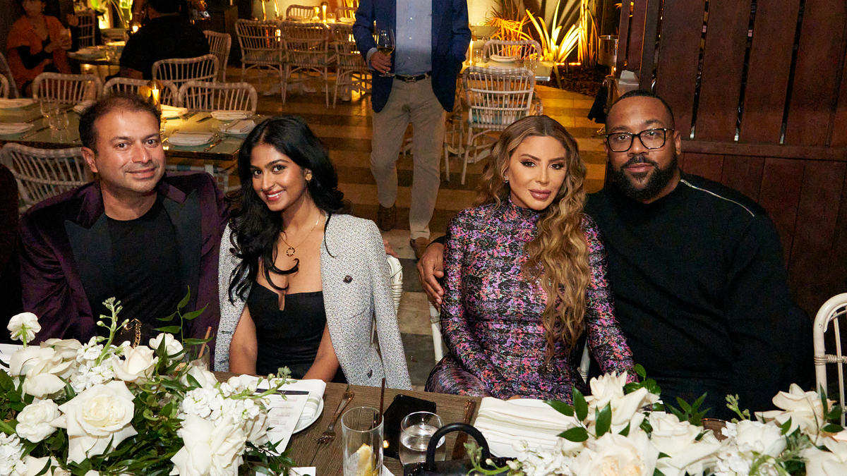 Four people seated at a dinner table with white floral centerpieces, smiling. Two women, one in tweed and the other in a patterned dress