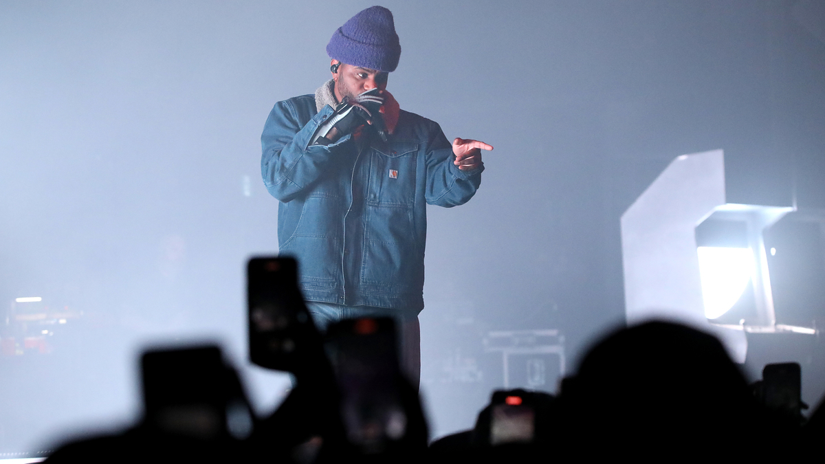 Musician in denim jacket and beanie performs on stage, audience with raised phones in foreground