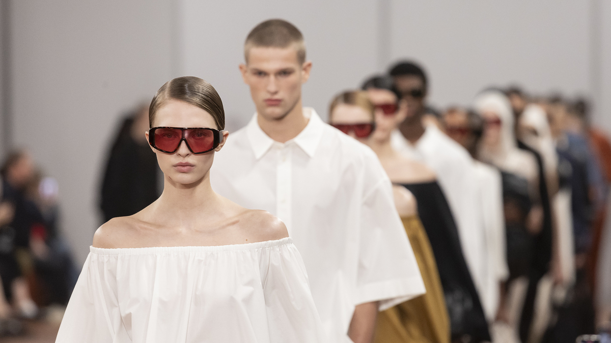 Models in a fashion show wearing minimalist outfits, with a focus on a female model in a white off-the-shoulder top