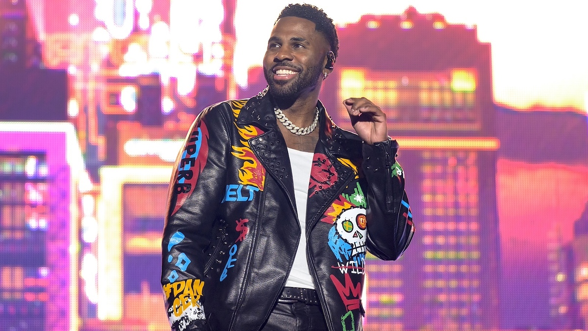Man smiling on stage wearing a decorated leather jacket with graphic designs and a necklace