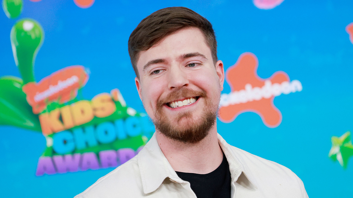 Smiling man at the Kids' Choice Awards with a logo backdrop, wearing a casual beige jacket