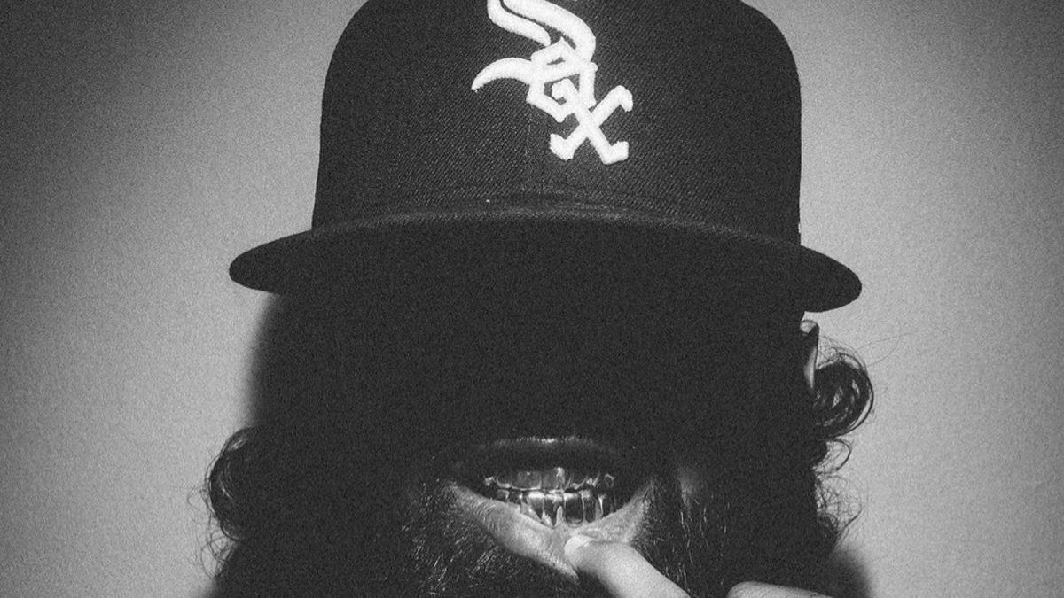 Close-up of a person with a beard wearing a baseball cap, with their finger on their lips. Includes "Parental Advisory" label