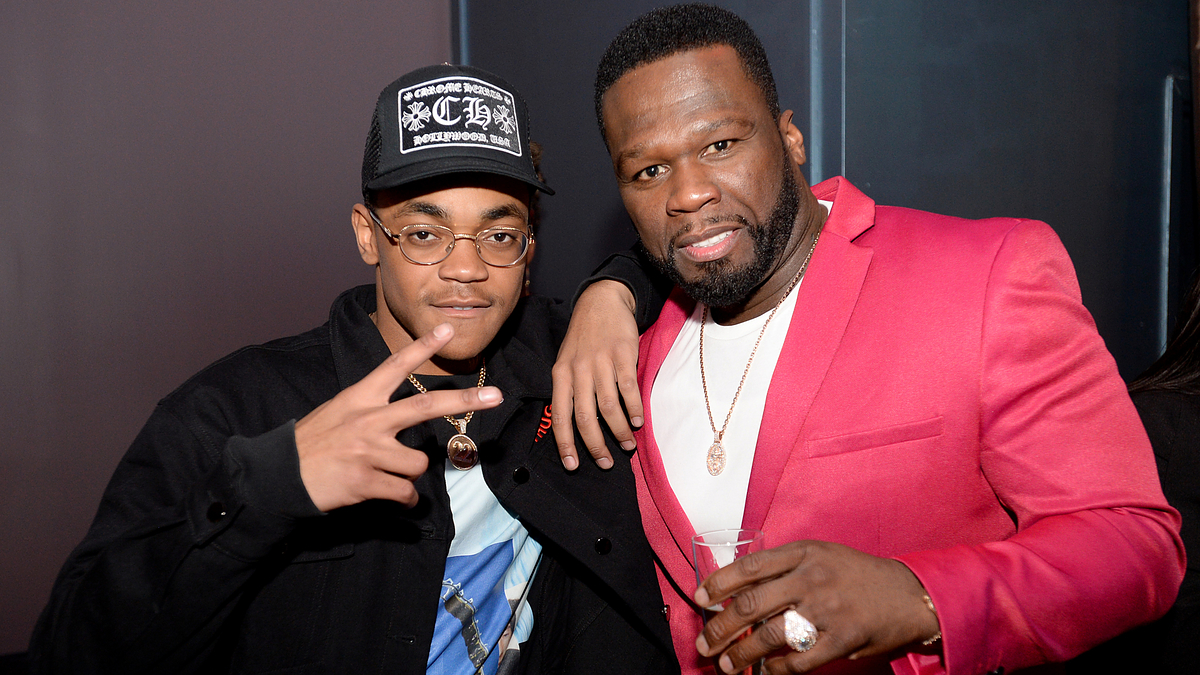 Two men posing for the photo, one in a red blazer and another wearing a cap and glasses