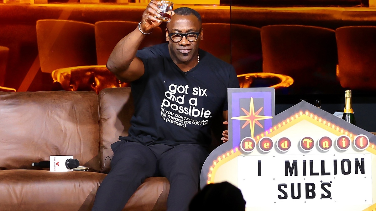 Person in black t-shirt sitting, raising glass, sign with text "1 MILLION SUBS" nearby