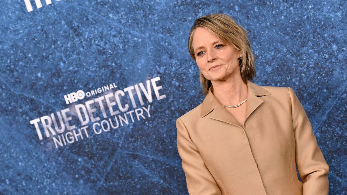 Jodie Foster standing in front of a backdrop with "HBO Original True Detective Night Country" text, wearing a stylish beige suit