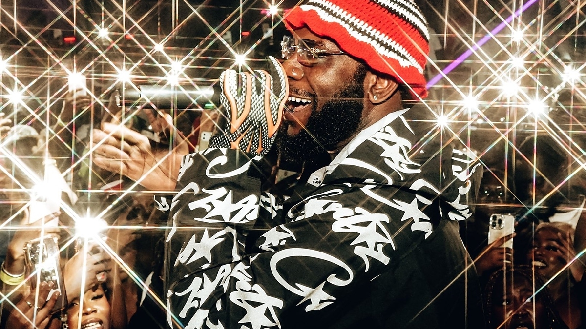 Gucci Mane is performing on stage wearing a patterned jacket and a colorful striped beanie, with fans in the crowd capturing photos with their phones