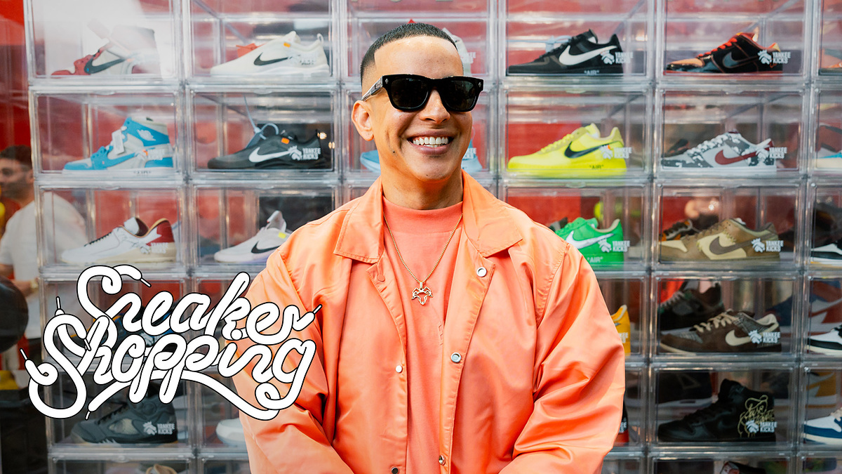 Daddy Yankee smiling while wearing sunglasses in front of a wall filled with sneakers.