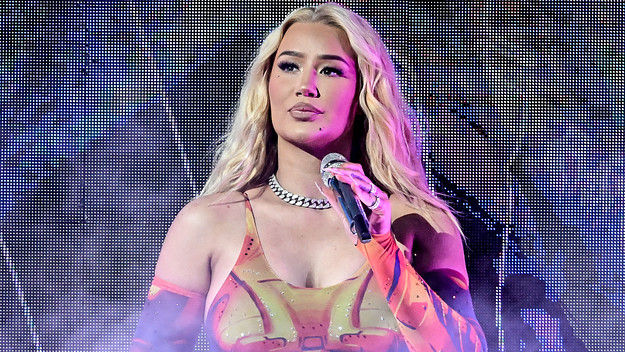 Iggy Azalea performing on stage, holding a microphone. She has long blonde hair and is wearing a colorful outfit.