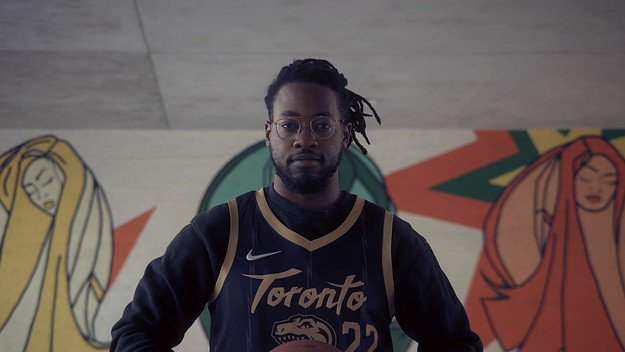 Photo of Gordon Rowe Centred, wearing a black and gold Raptors jersey and holding a basketball. Behind him is a colourful mural featuring line-drawn women.