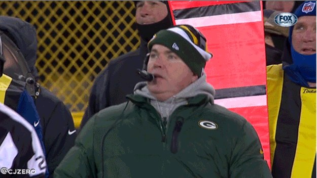 A football coach in a Green Bay Packers jacket and beanie, with a headset, stands on the sidelines during a game.