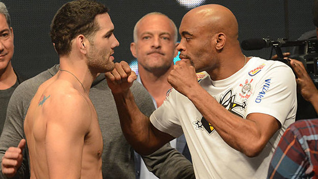 Two fighters face off at a weigh-in, with one shirtless and the other in a white shirt, surrounded by people and cameras.