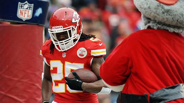 A Kansas City Chiefs player in a red jersey runs with a football, facing a person in a Santa costume. NFL logo visible.