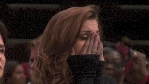 A woman with long brown hair covers her face with her hands, appearing emotional. People are in the background.