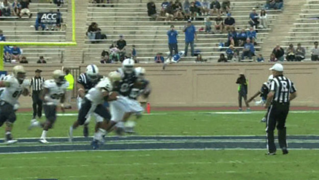 A college football game in progress, with players in blue and white uniforms on the field, and a referee observing nearby.