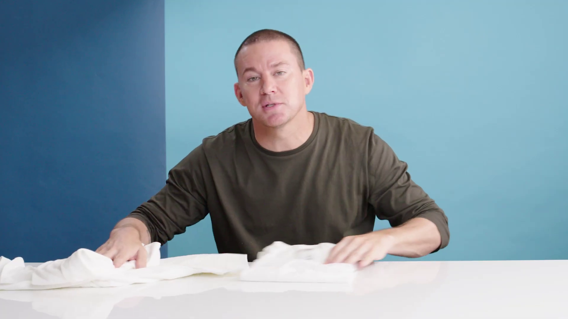 Channing Tatum sitting at a table with a small pile of white t-shirts