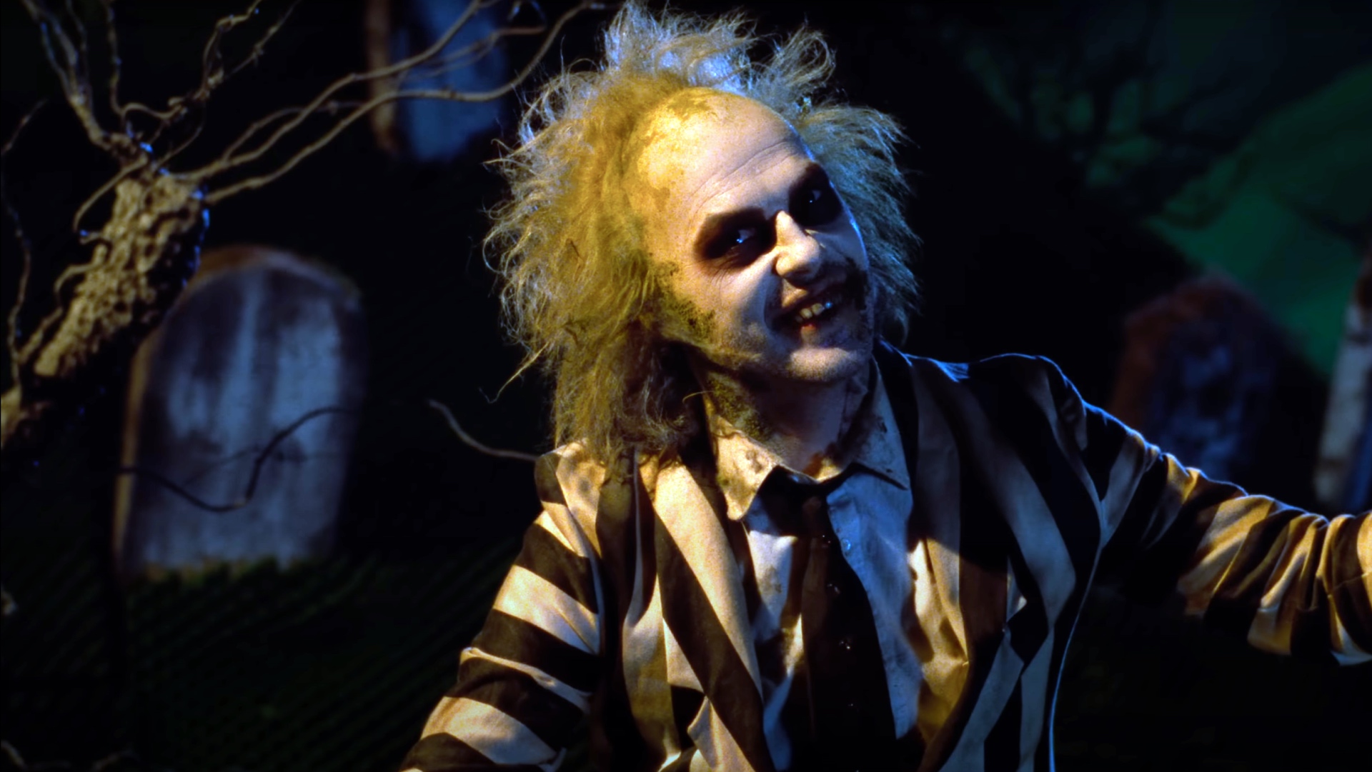 Beetlejuice (Michael Keaton) poses against a cemetery with a smile on his face and his arms outstretched.