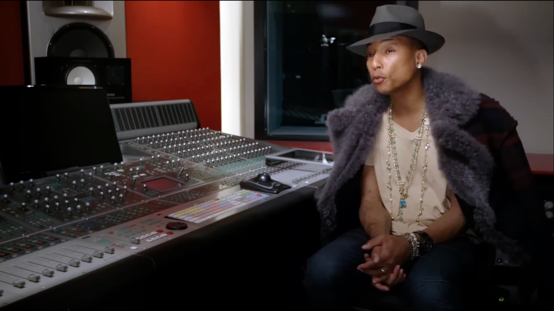 Pharrell Williams sits inside a recording booth in a fur coat and fedora.