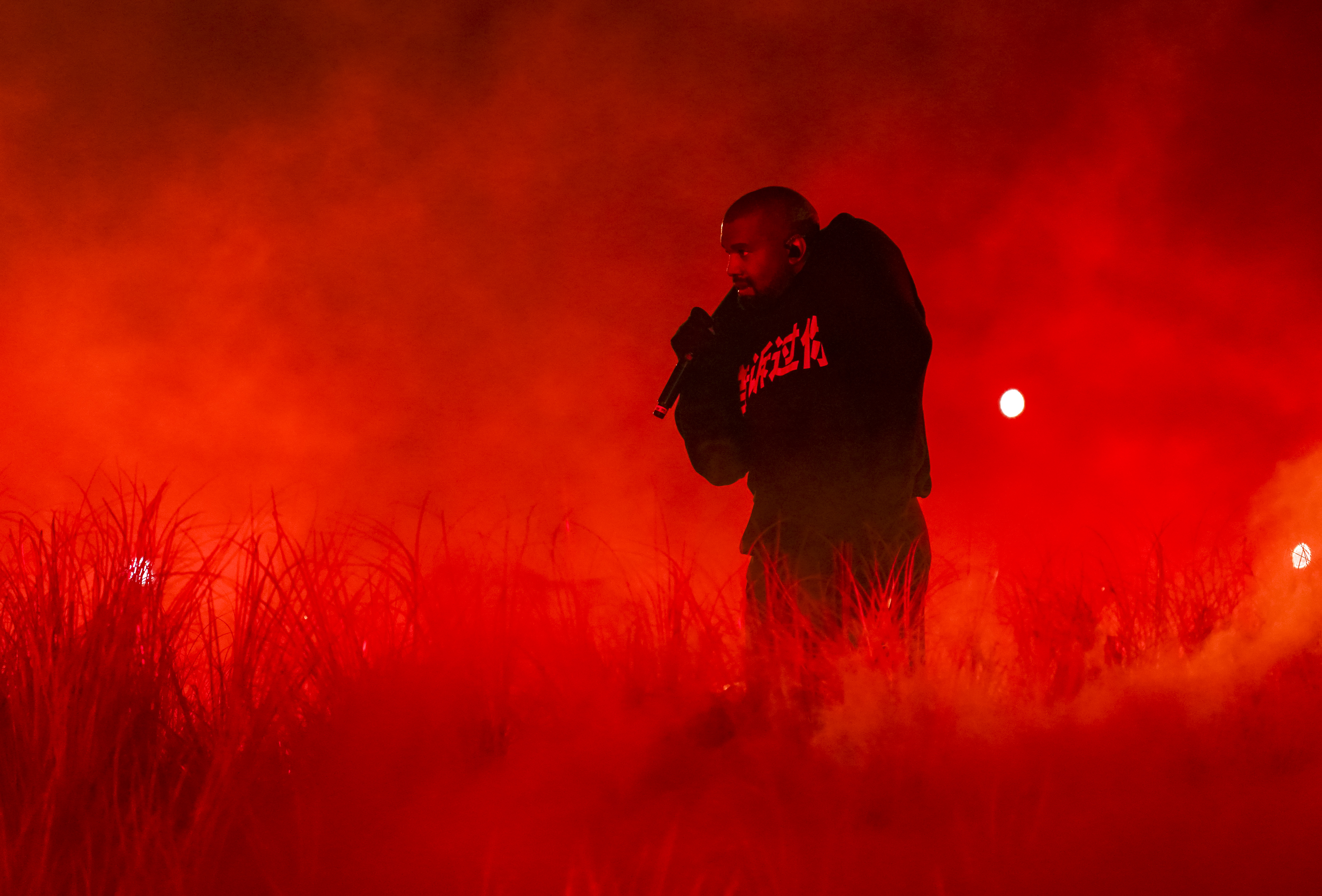 Kanye West performing on stage, surrounded by red smoke and dramatic lighting, wearing a dark hoodie and holding a microphone.