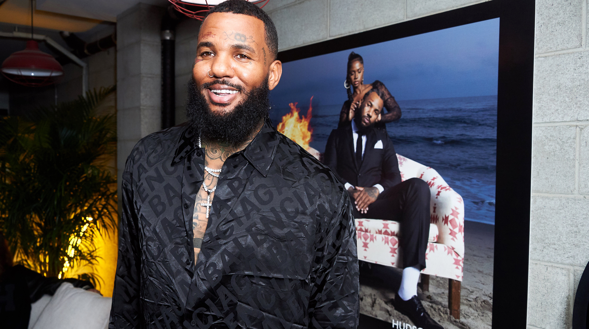 The Game, wearing a black shirt, stands smiling in front of a large photo of himself seated on a beach.