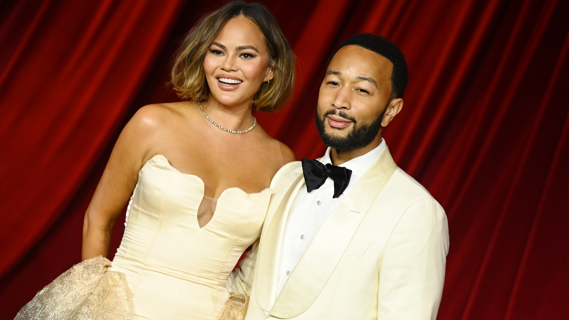 Chrissy Teigen and John Legend pose together on a red carpet. Chrissy wears a strapless gown, and John is in a white tuxedo.