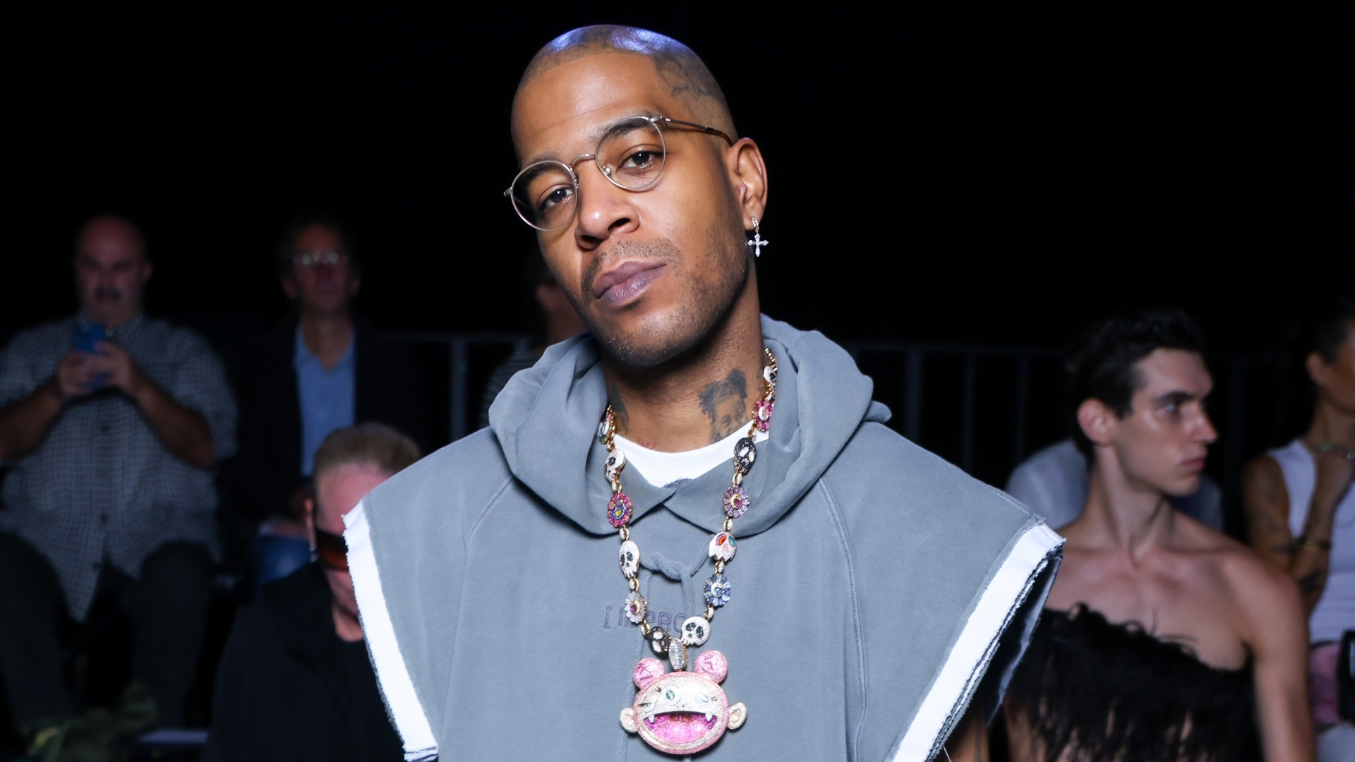 Kid Cudi wearing glasses and a unique necklace, standing in a fashion show audience.