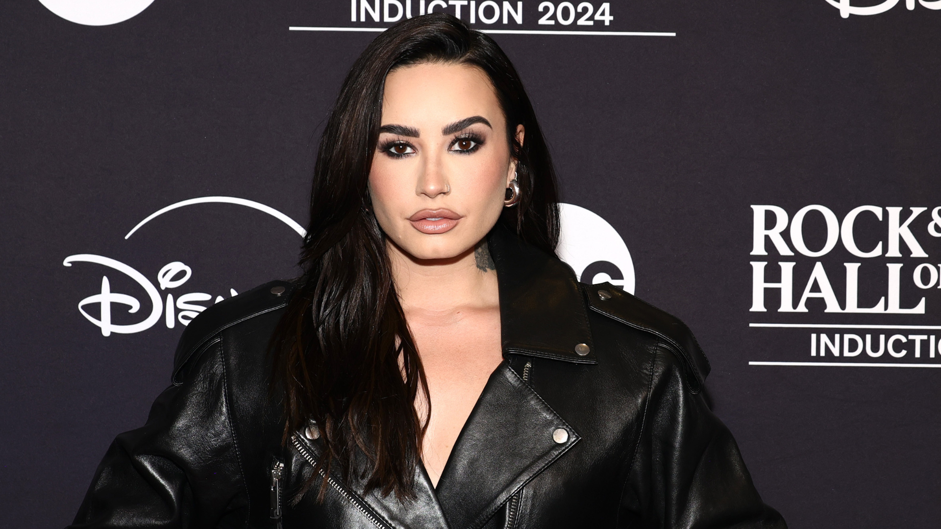 Demi Lovato at the Rock & Roll Hall of Fame Induction 2024 event, wearing a black leather jacket, standing against a branded backdrop.