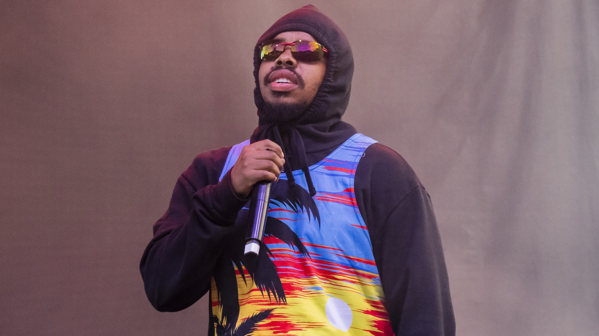 Earl Sweatshirt wearing a colorful hoodie and sunglasses holds a microphone on stage.