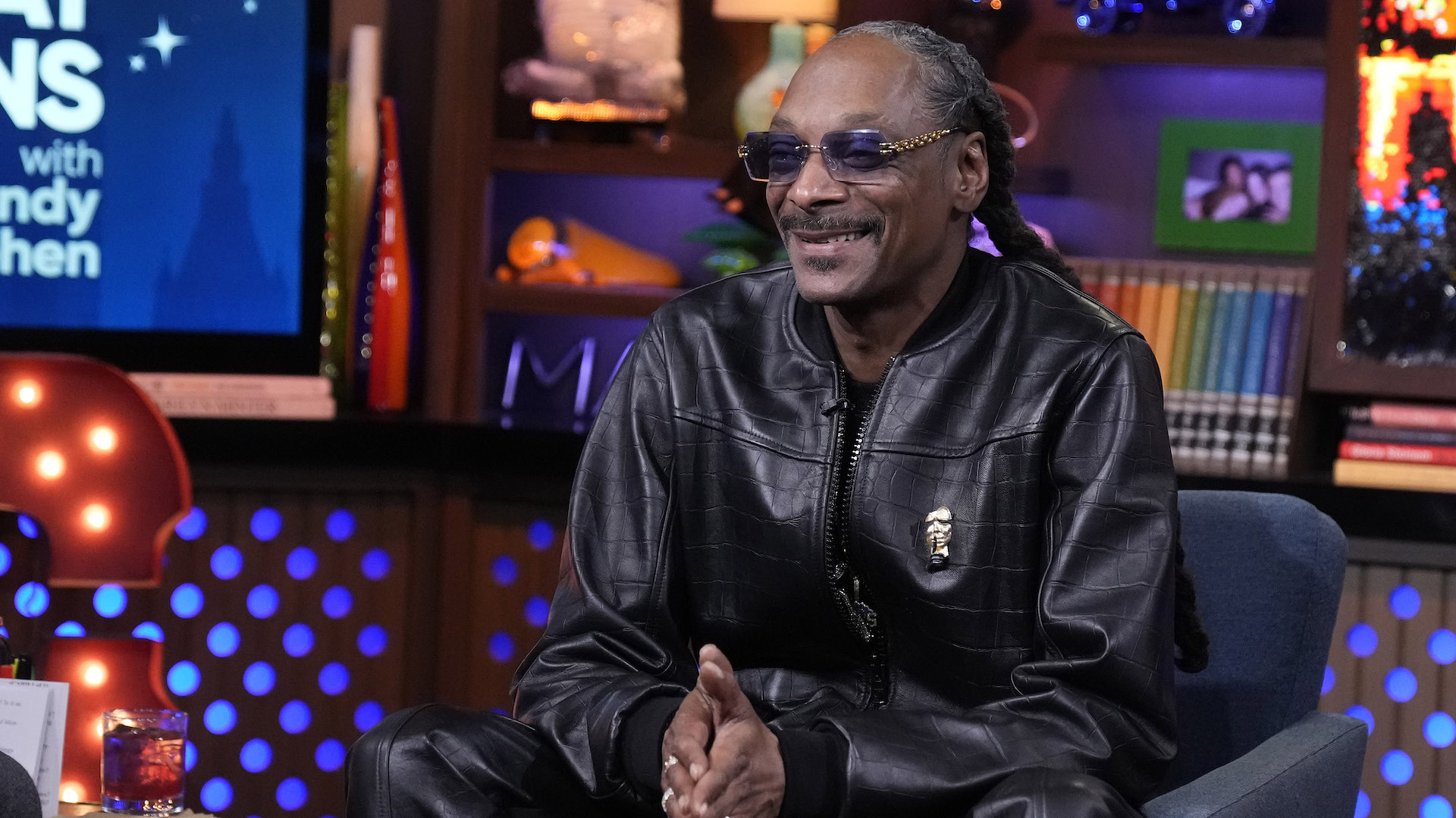 Snoop Dogg, wearing sunglasses and a black leather outfit, sits smiling on a talk show set with colorful decor in the background.