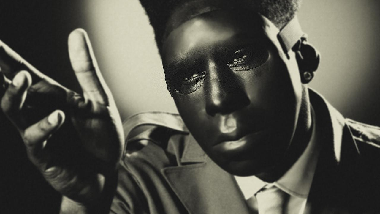 A person in a trench coat and tie with a shadowed face, reaching out with one hand in a dramatic, noir-style image.