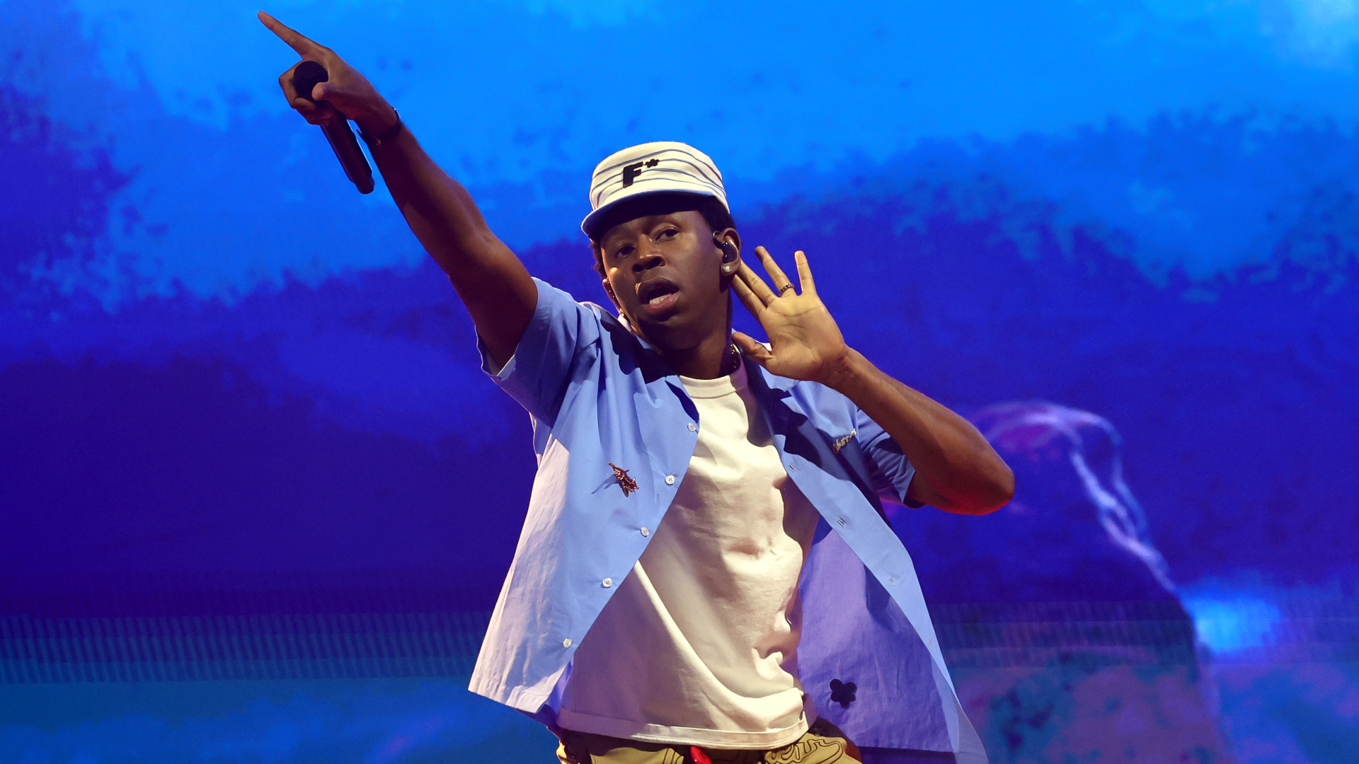 Tyler, the Creator performing on stage, wearing a blue shirt and striped hat, holding a microphone, with a vibrant blue background.
