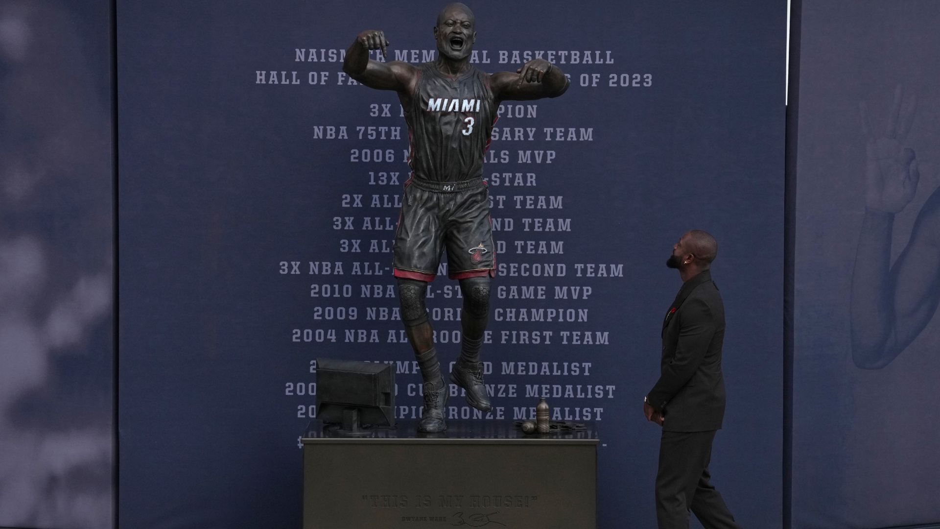 A man in a suit looks at a statue of a basketball player wearing a Miami uniform, with career achievements listed in the background.