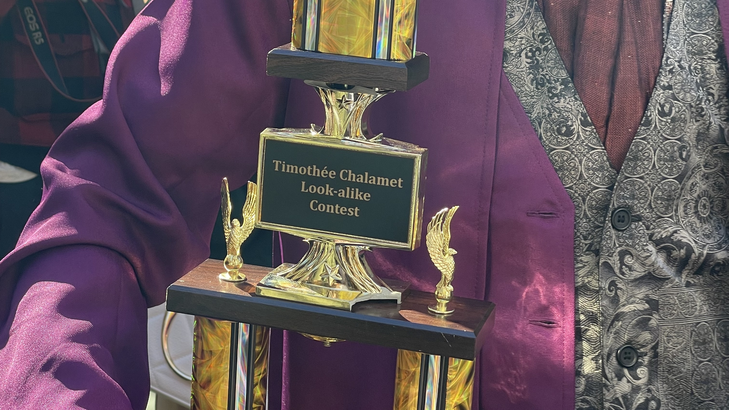 A person in a purple suit holds a trophy labeled "Timothée Chalamet Look-alike Contest."