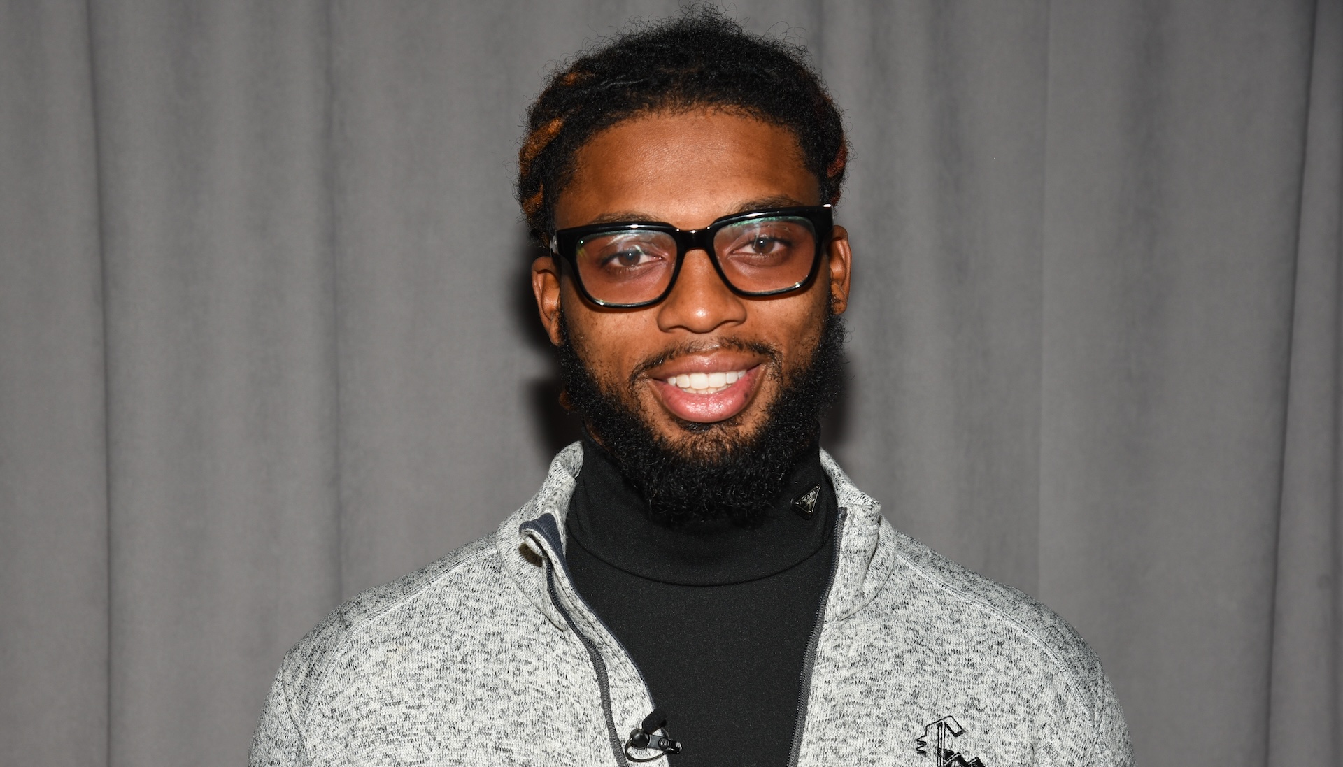 Damar Hamlin with glasses and a beard, wearing a gray zip-up jacket over a black shirt, stands in front of a gray curtain.