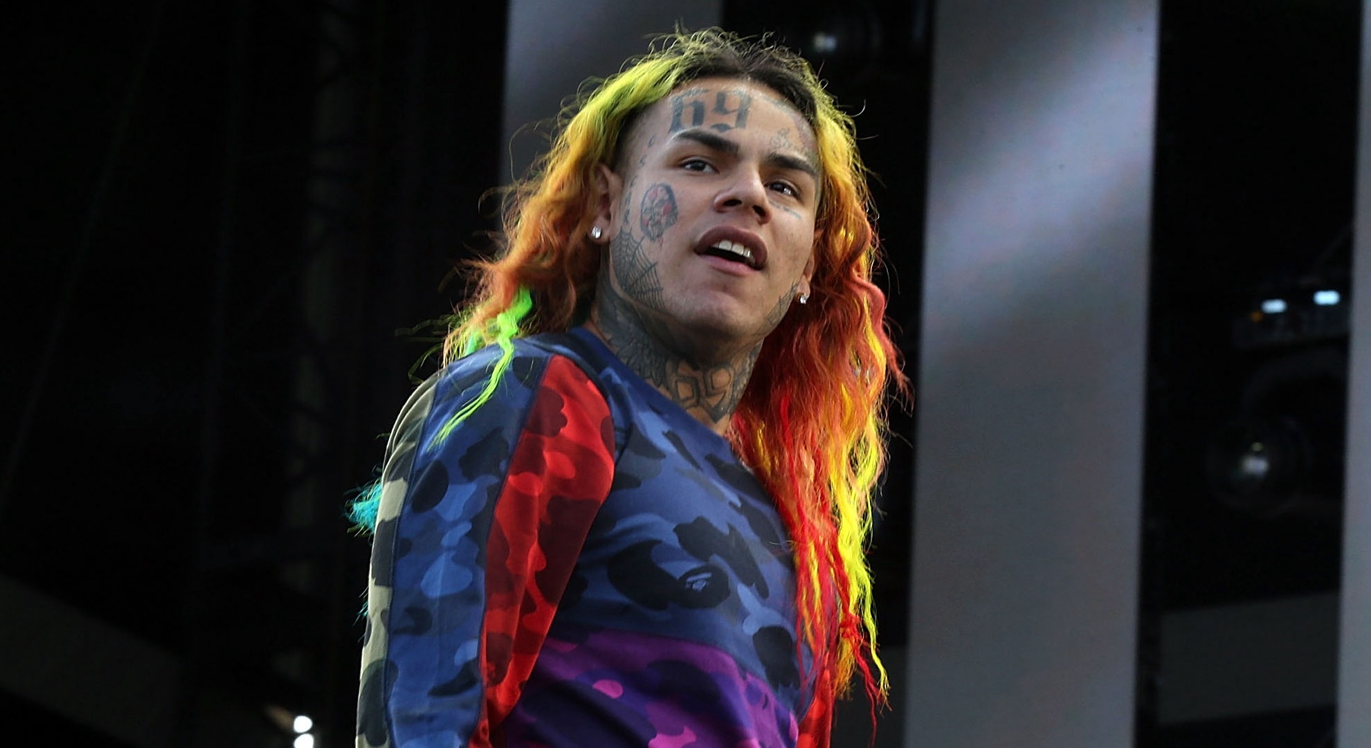 Rapper Tekashi 6ix9ine with colorful hair and face tattoos, wearing a multicolored shirt, performing on stage.