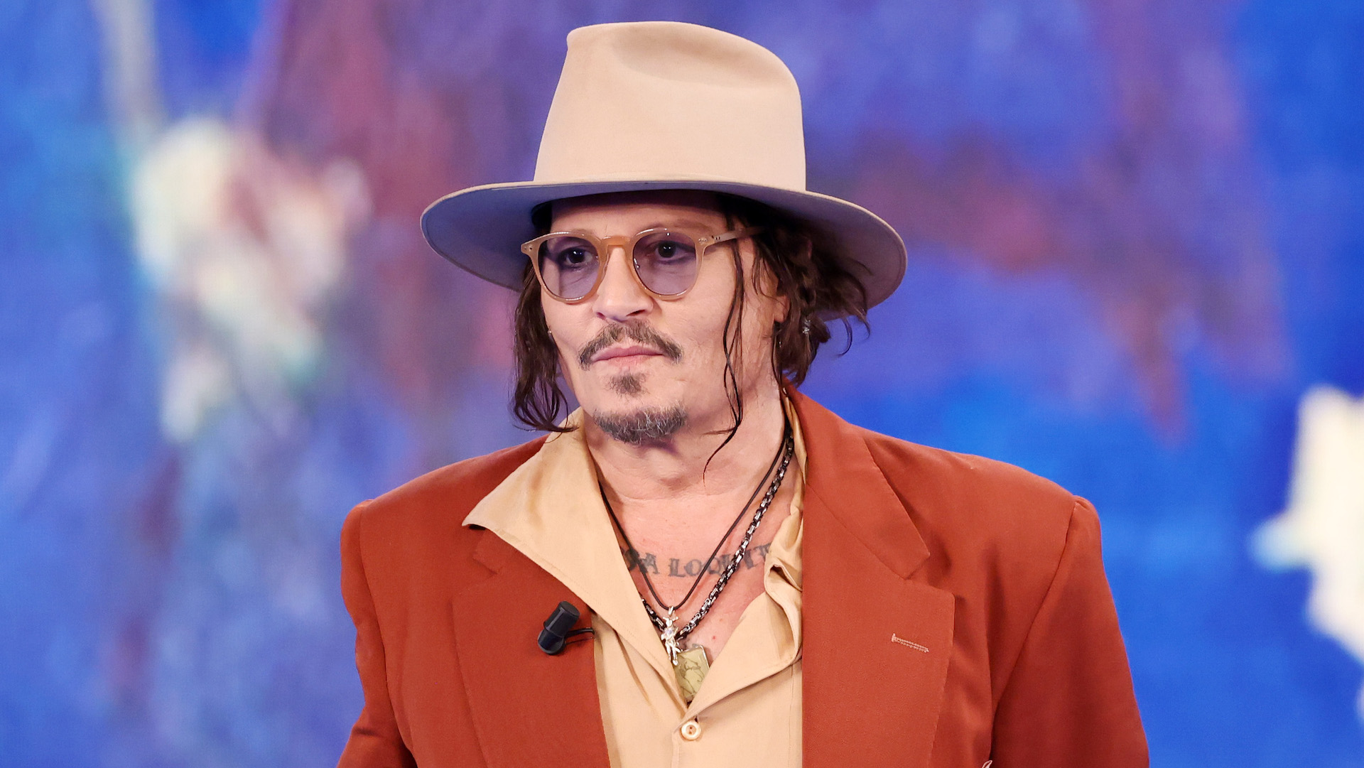 Johnny Depp wearing a tan hat, sunglasses, and a rust-colored suit jacket over a beige shirt, standing against a blue background.