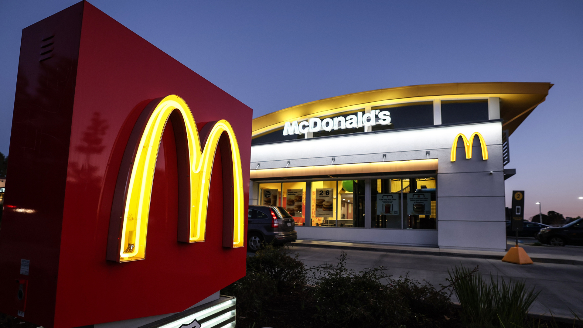 A McDonald's restaurant at dusk with illuminated golden arches and a large red sign in the foreground.