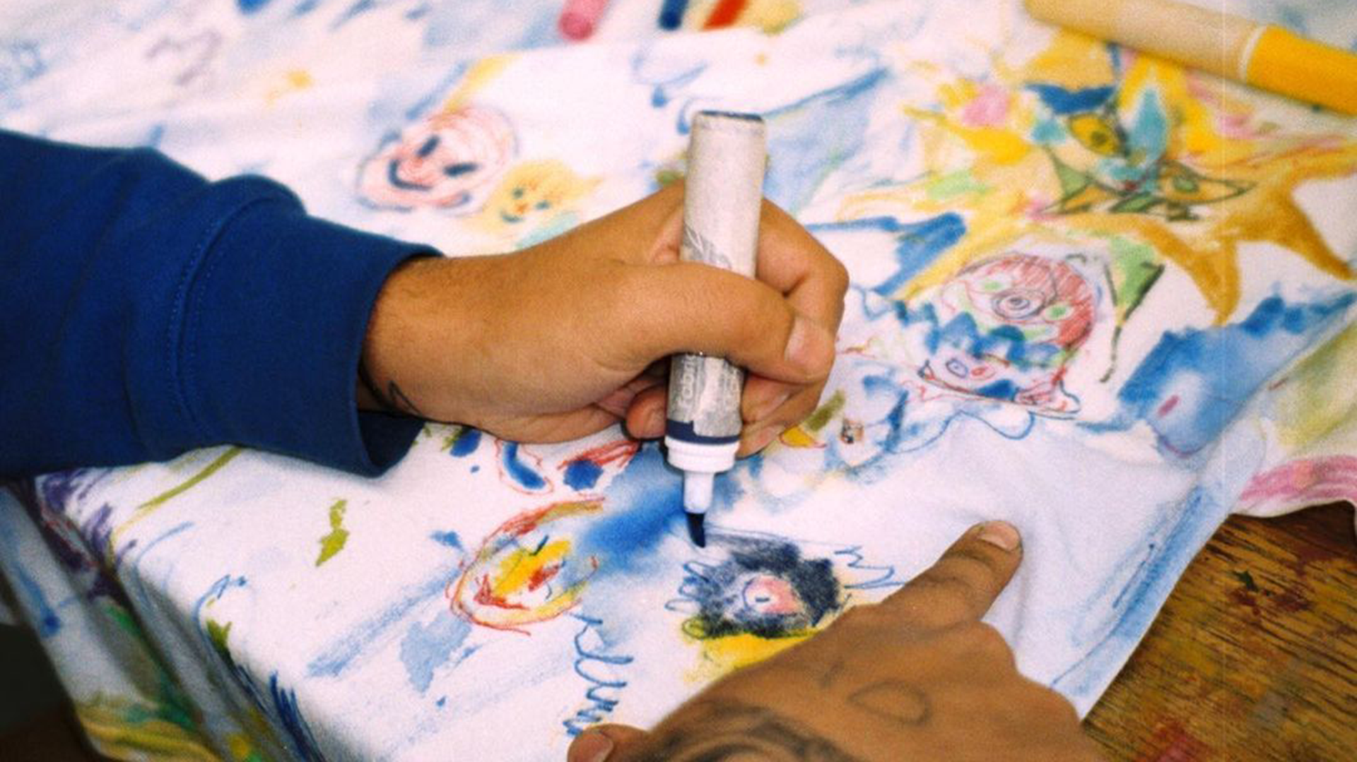 A person drawing colorful cartoon characters on fabric with a marker, with visible tattoos on their hand. Tony tafuro