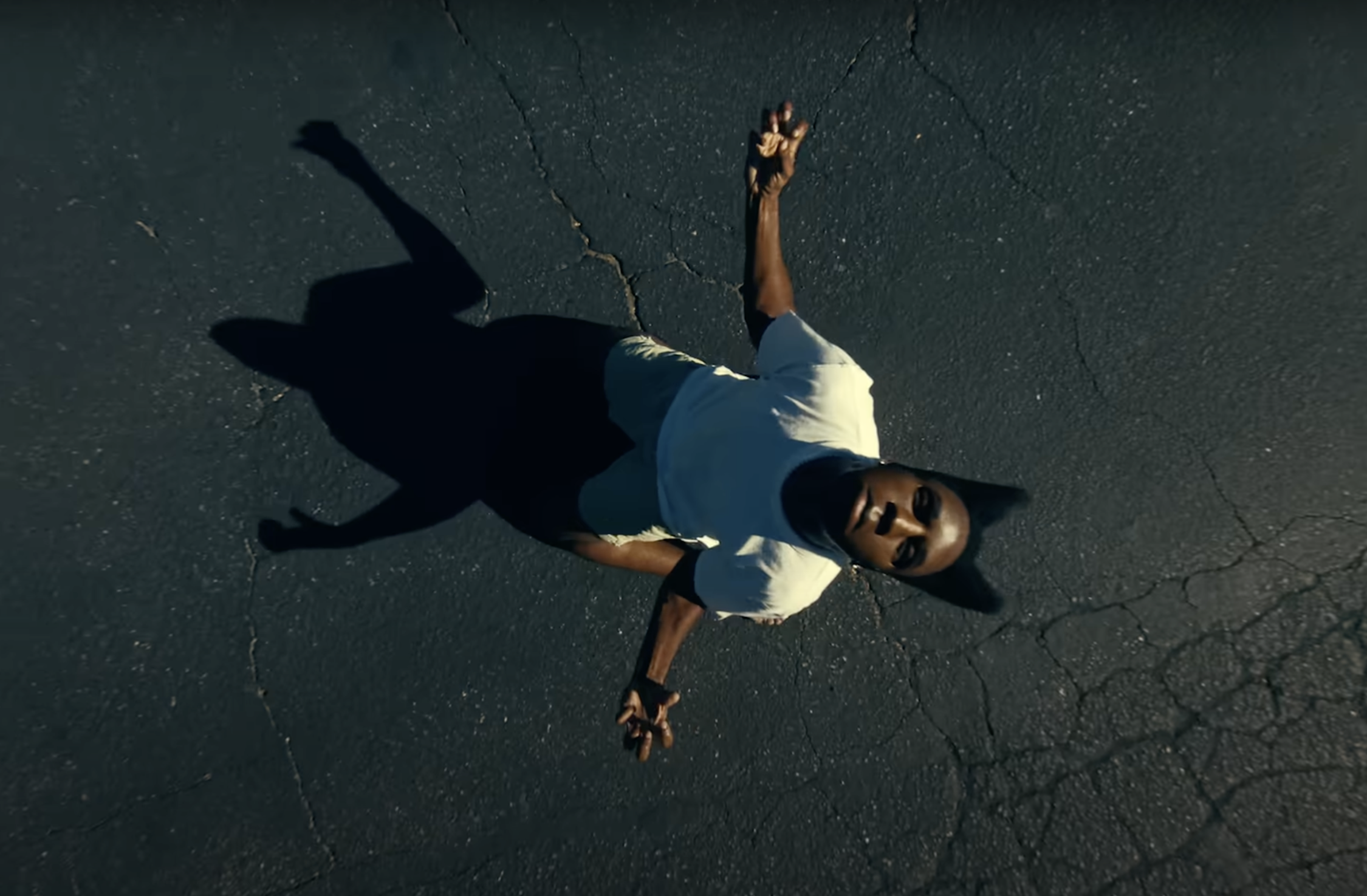 Aerial view of Tyler, the Creator in a white shirt standing on cracked pavement, casting a long shadow.