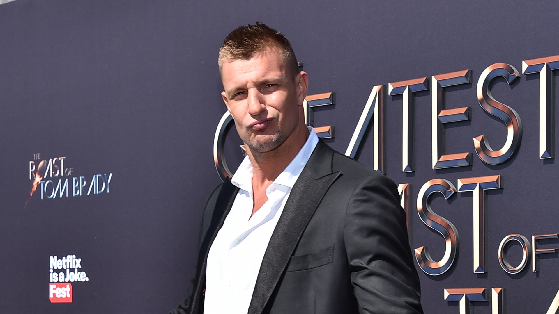 Rob Gronkowski at a Netflix event, wearing a black suit and white shirt, standing in front of a backdrop with event branding.