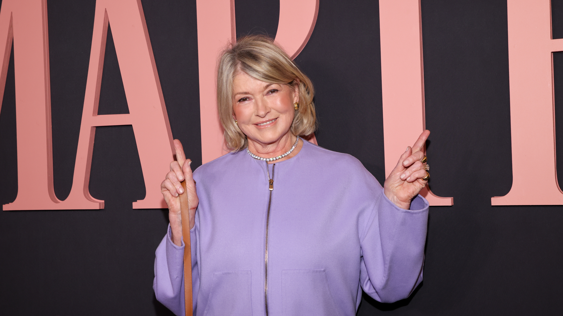 Martha Stewart in a lavender outfit, smiling and posing with finger guns against a backdrop with large letters.