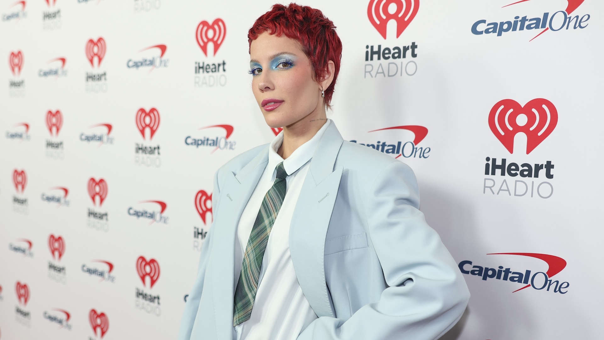 Halsey wears a light blue blazer and green plaid tie with red pixie haircut and blue eyeshadow on a red carpet.