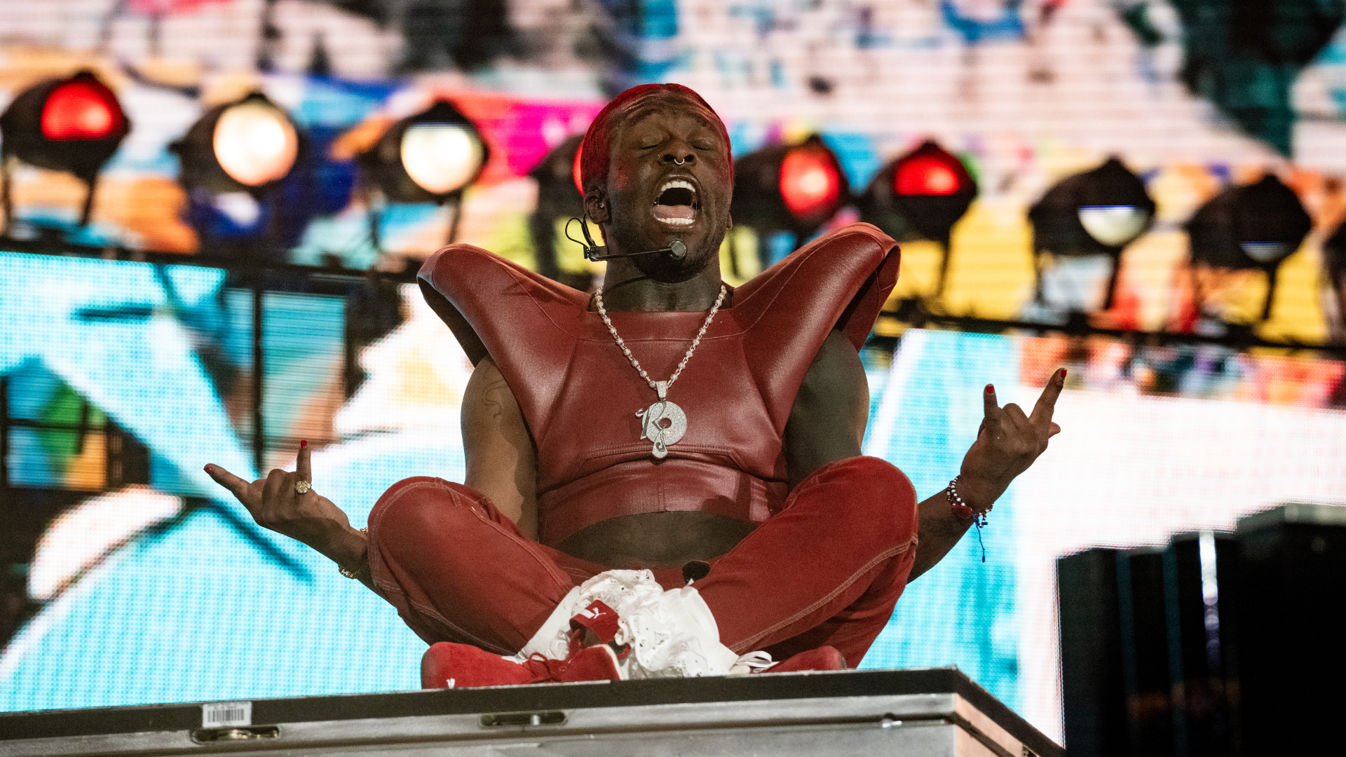 A performer in a red outfit and headband sits cross-legged on stage, passionately singing. Colorful lights and a vibrant backdrop enhance the scene.